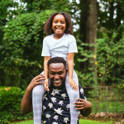 A man stands outdoors with a child sitting on his shoulders, both smiling. They are in a lush, green garden with trees in the background.